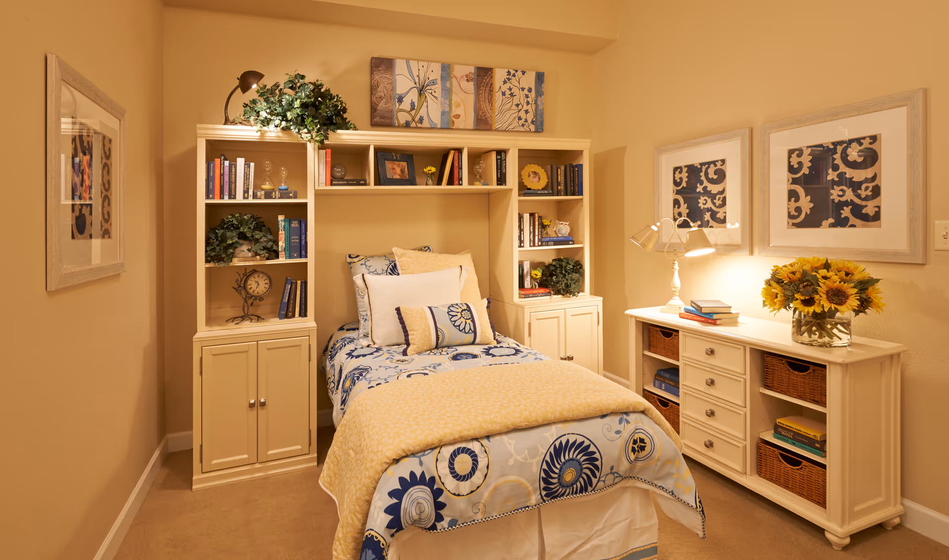 A cozy bedroom with a single bed featuring a blue and yellow floral patterned bedspread and multiple pillows. The bed is flanked by built-in shelves filled with books, plants, and decorative items. A white dresser with wicker baskets and a vase of sunflowers sits against the right wall, illuminated by a table lamp. The walls are painted a warm beige and decorated with framed artwork.