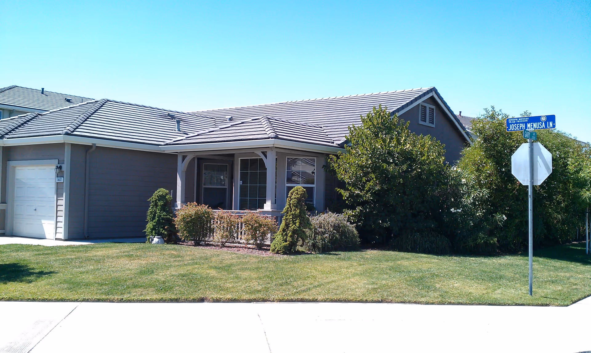Single-story residential building with a gray exterior and tiled roof, surrounded by a well-maintained lawn and various bushes. A street sign at the corner reads Joseph Menusa Ln.