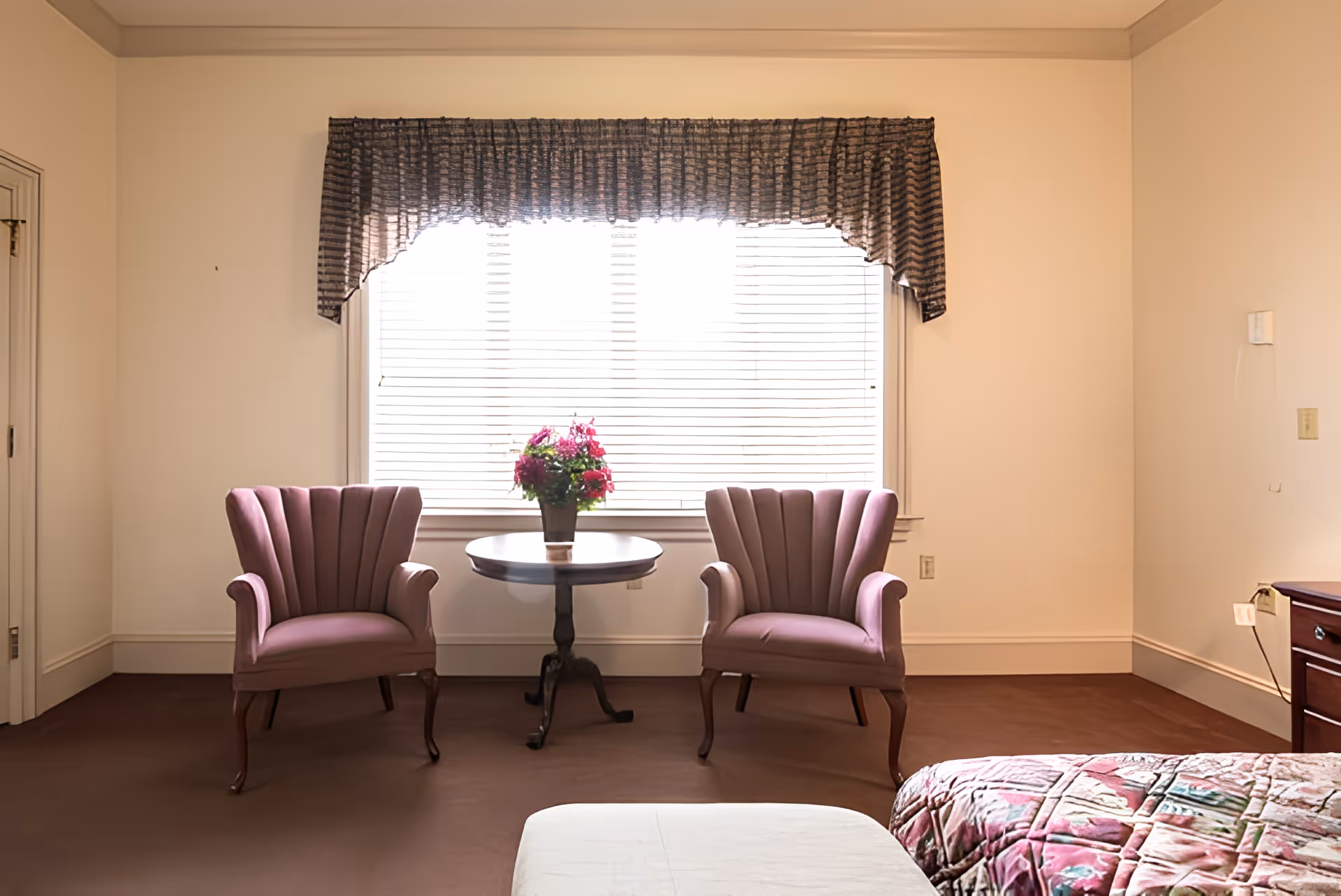 A cozy room with two mauve upholstered armchairs positioned on either side of a small round wooden table with a flower pot containing pink and red flowers. Behind the chairs is a large window with white blinds and a dark valance. Part of a bed with a colorful quilt is visible on the right side, and a wooden nightstand with a lamp is also partially visible.