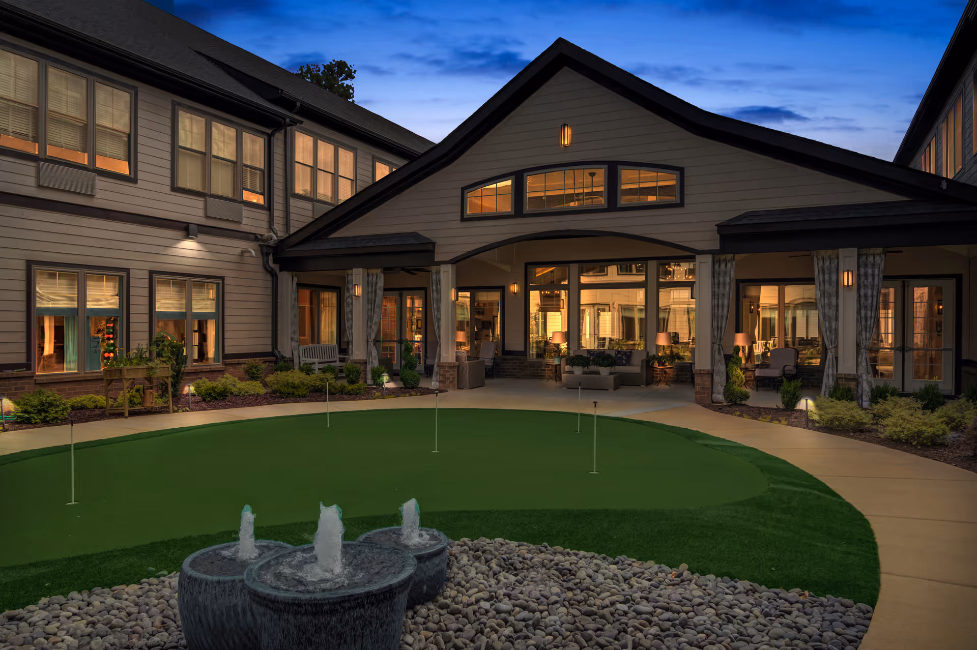 Evening view of a senior living facility courtyard at Oaks at Douglasville featuring a small putting green with four flags, a three-tiered water fountain surrounded by pebbles, and a covered patio area with seating and warm lighting inside the building.