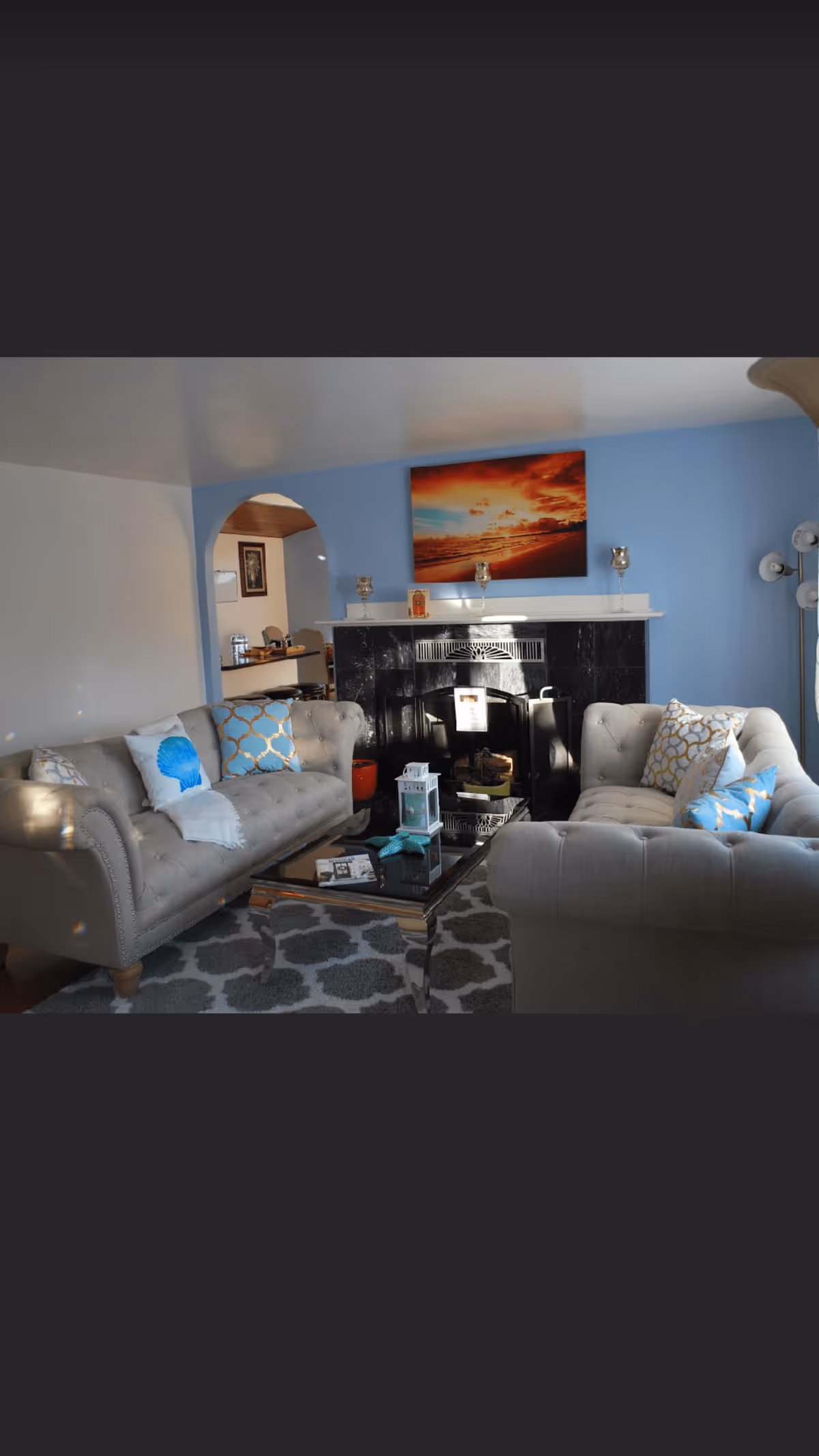 Cozy living room with two tufted sofas facing a glass coffee table and a fireplace beneath a sunset painting on a blue accent wall.