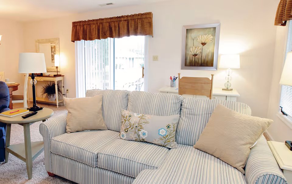 A cozy living room with a striped sofa adorned with three cushions, a side table with a lamp and remote controls, a wooden chair with a desk behind it, a framed floral painting on the wall, and a sliding glass door with vertical blinds and a brown valance.