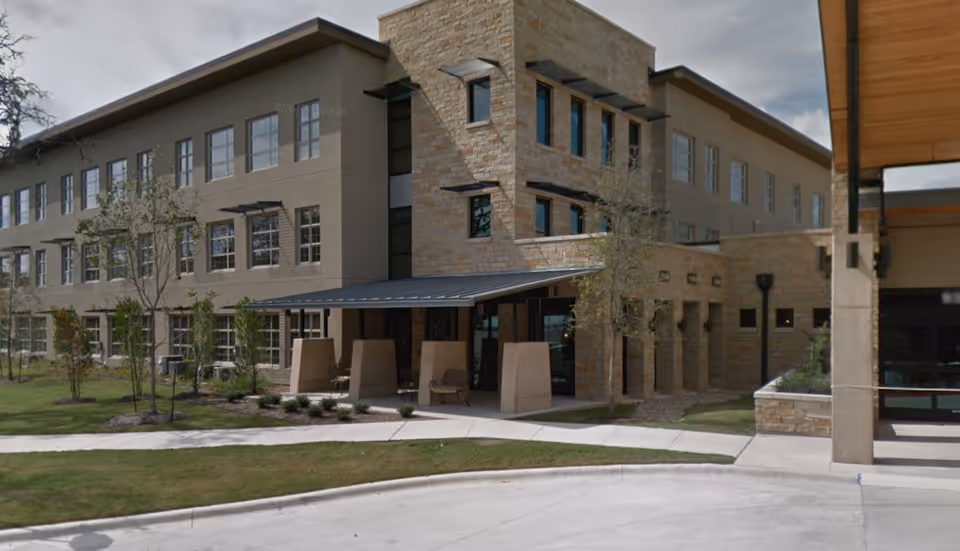 Exterior view of a modern three-story building with a combination of stone and beige stucco walls, large windows, and a covered entrance area. There are small trees and landscaped bushes around the building, with a curved driveway in front.