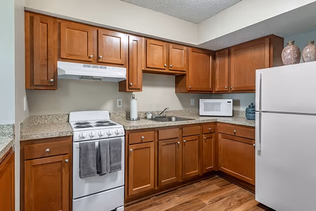 A kitchen with wooden cabinets, a white stove with two gray towels hanging on the handle, a white microwave on the countertop, a white refrigerator, and a sink with a paper towel holder and some small containers on the counter. The floor is wooden and the walls are light-colored.