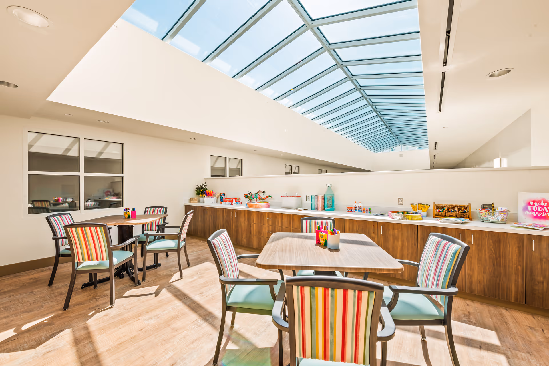 Bright room with a long skylight ceiling, featuring several tables and chairs with colorful striped upholstery. Along one wall, there is a countertop with various craft supplies and decorations. The room has wooden flooring and large windows on one side.