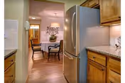 View of a kitchen with wooden cabinets and granite countertops, featuring a stainless steel refrigerator. Beyond the kitchen is a dining area with a round table, four chairs, a hanging light fixture, and a decorative plant on the table.