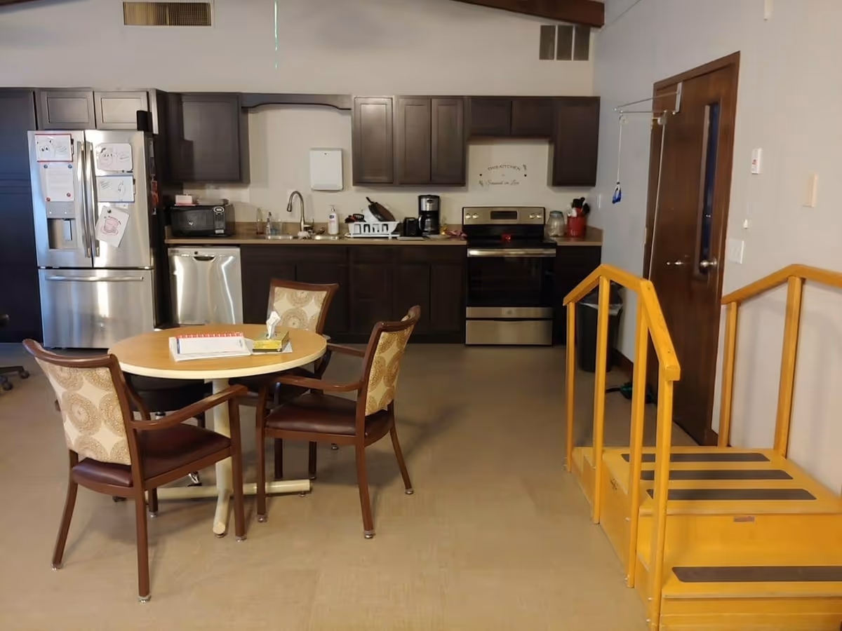 Communal kitchen and dining area with a round table and four chairs, stainless steel appliances and dark cabinets, and a yellow safety step/railing on the right.