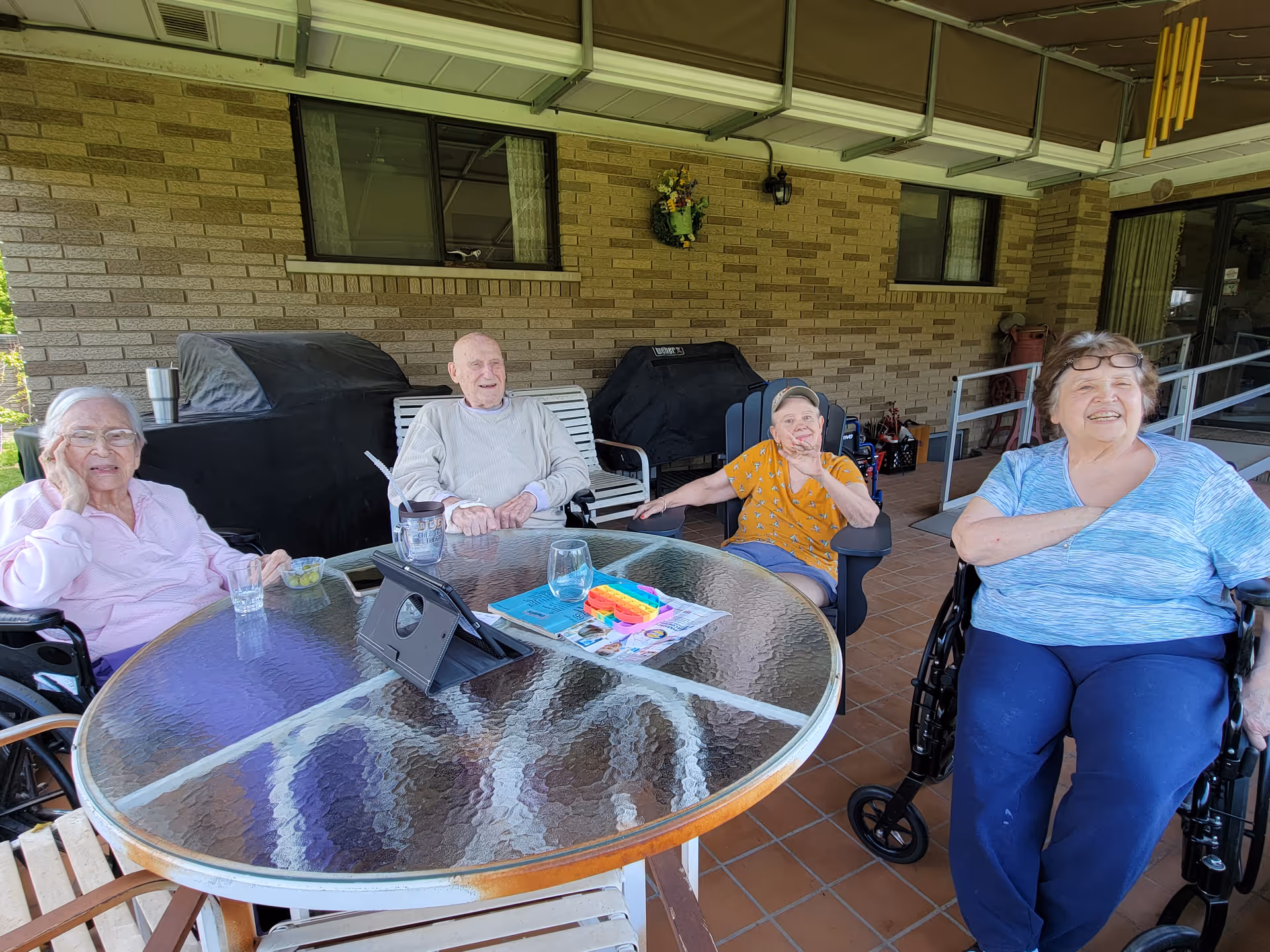 Four elderly individuals sitting around a glass outdoor table on a covered patio. Two women in wheelchairs are on the left and right sides, a man in a light sweater is seated on a bench, and another woman in a yellow shirt and cap is seated in a chair. The patio has a brick wall, two covered grills, and a wind chime hanging from the ceiling.