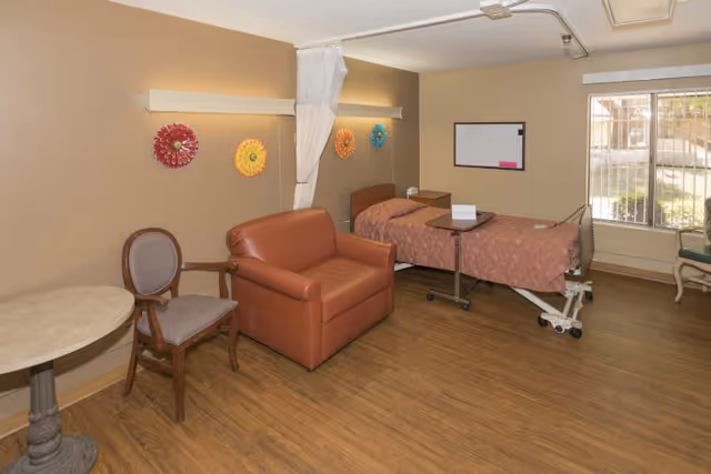 A rehabilitation center room with a hospital bed covered in a pink patterned blanket, a brown leather loveseat, a wooden chair, a small round table, and colorful decorative wall hangings. There is a window with bars and a whiteboard on the wall.