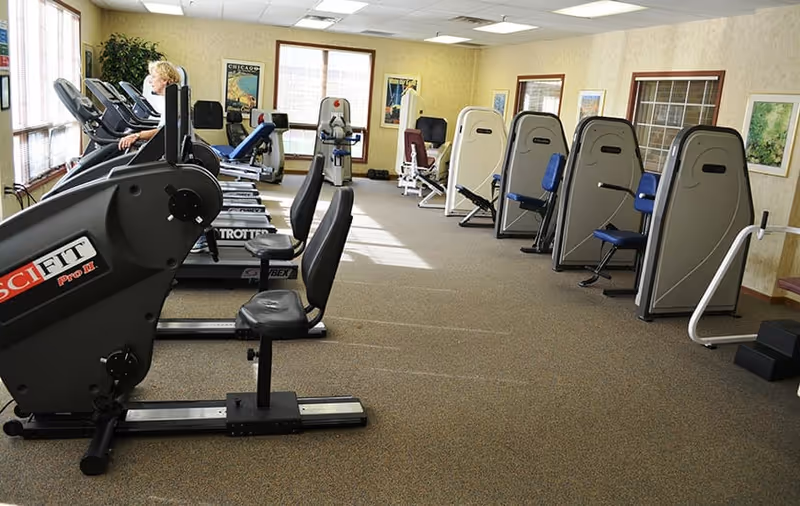 A fitness room with various exercise machines including stationary bikes and weight machines. A person is using one of the machines near the windows. The room has carpeted floors, beige walls, and several framed pictures on the walls.