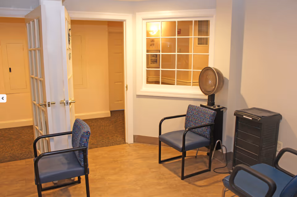 A small waiting area with three blue cushioned chairs with black metal arms, a vintage hair dryer on a stand, and a black rolling cart. The room has light-colored walls, a wooden floor, and a window with white trim looking into another room. There is an open door leading to a carpeted hallway.