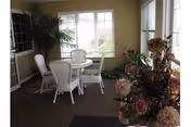 A sunlit indoor seating area with a round glass table and four white wicker chairs. Large windows allow natural light to fill the space, and there are plants and floral arrangements adding decoration.