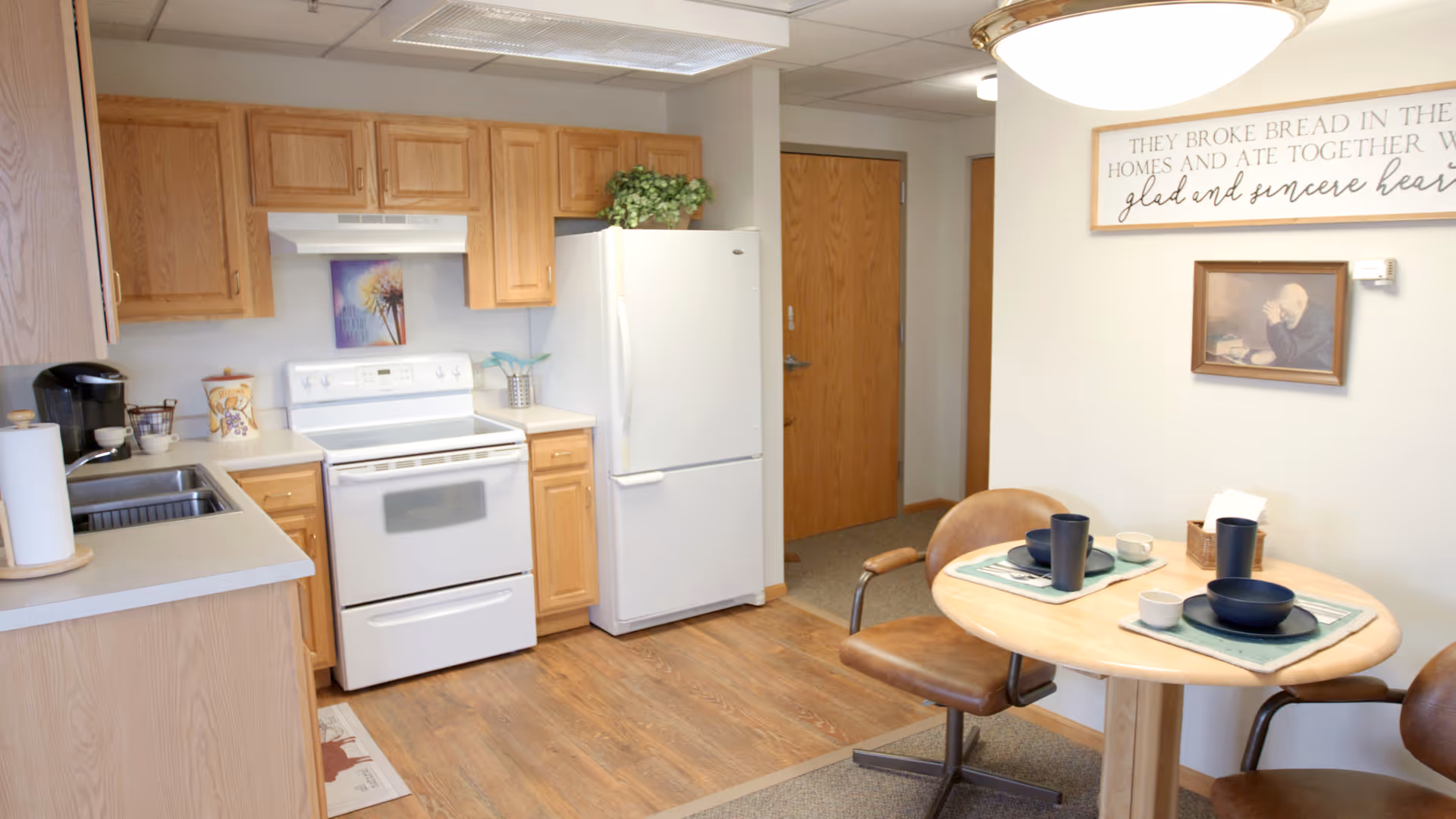 A small kitchen and dining area with light wood cabinets, a white stove and refrigerator, and a round wooden table set for two with brown chairs. There is a framed picture and a decorative sign on the wall above the table. The floor is wood, and the room has a bright, clean appearance.