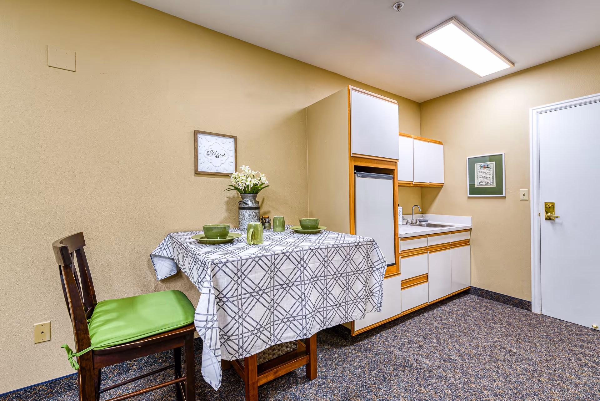 A small kitchenette area with a white and wood-trimmed cabinet, a mini refrigerator, a sink, and upper and lower storage cabinets. In front of the kitchenette is a small table covered with a white and gray checkered tablecloth, set with green dishes, bowls, and mugs. A wooden chair with a green cushion is placed next to the table. The walls are painted beige, and there is a framed picture and a decorative sign that says 'blessed' on the wall. The floor is carpeted in a blue and gray pattern, and a bright ceiling light illuminates the room.