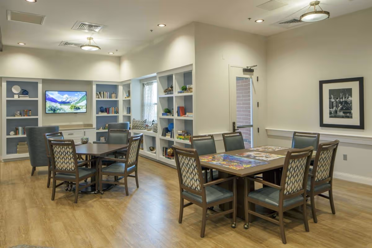 A well-lit common area with wooden flooring featuring two tables surrounded by chairs. One table has a puzzle in progress on it. The room has built-in shelves filled with books and decorative items, a window with a cushioned seating area, a door leading outside, and a wall-mounted TV displaying a scenic mountain landscape.