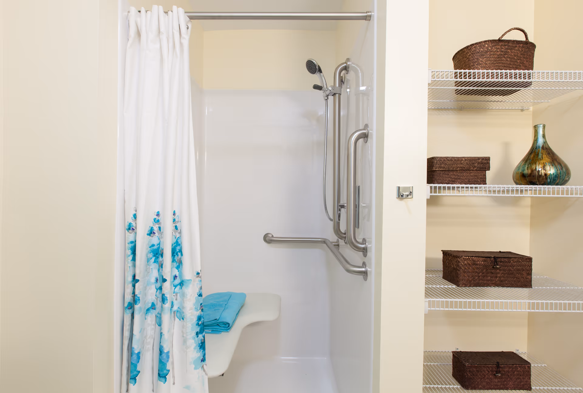 A walk-in shower with a white and blue floral curtain, stainless grab bars and a fold-down seat next to wire shelving holding wicker baskets and a decorative vase.