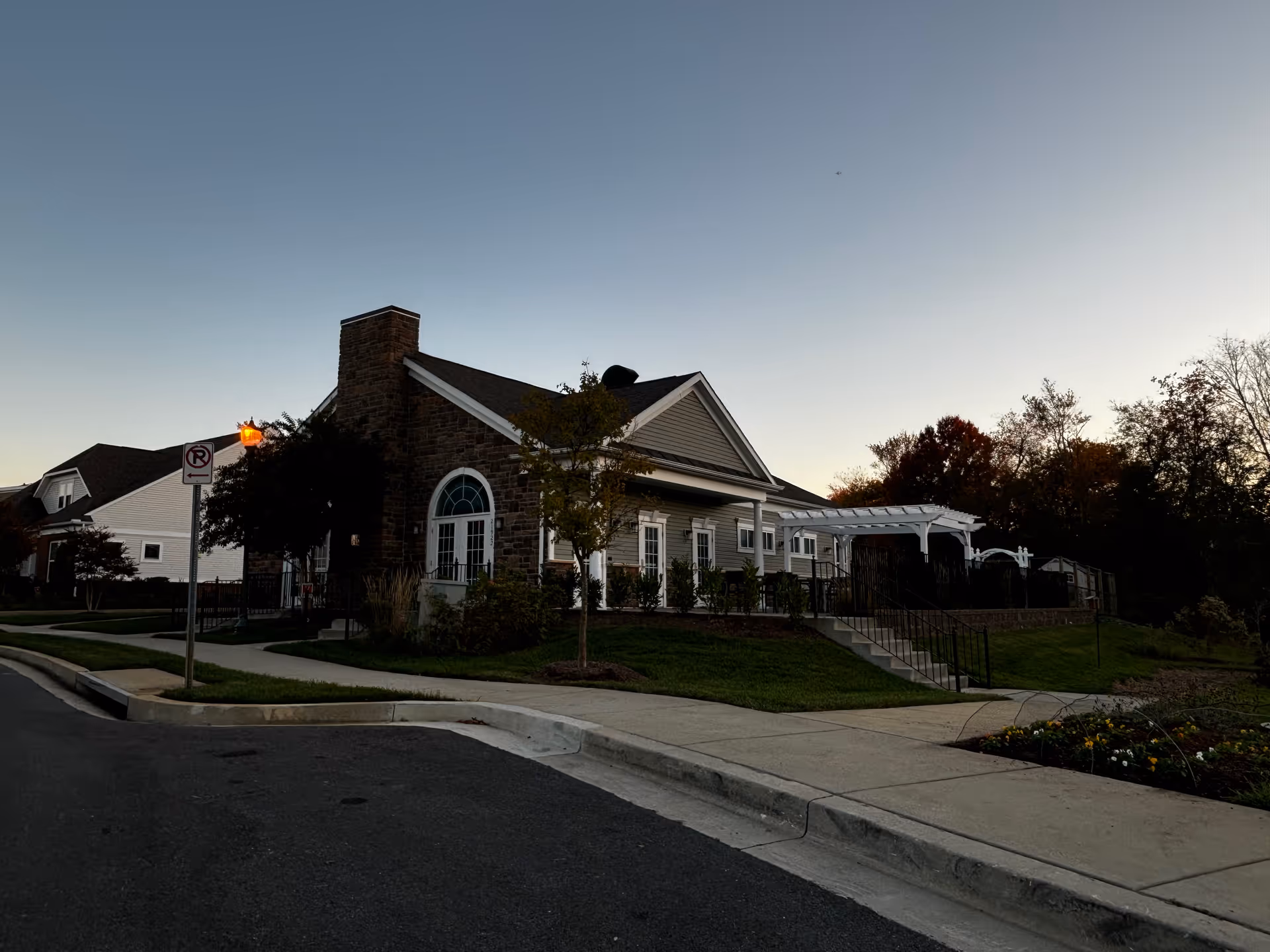 Exterior view of a single-story building with stone and siding facade at dusk, featuring a large arched window, a white pergola, stairs leading to a patio area, a sidewalk, and a street with a no parking sign and a lit streetlamp.