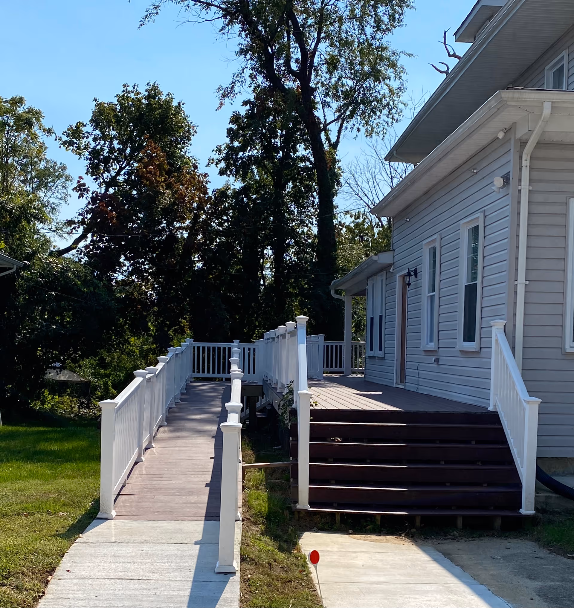 Exterior view of a building with a wooden ramp and stairs leading to a porch area, surrounded by green grass and trees under a clear blue sky.