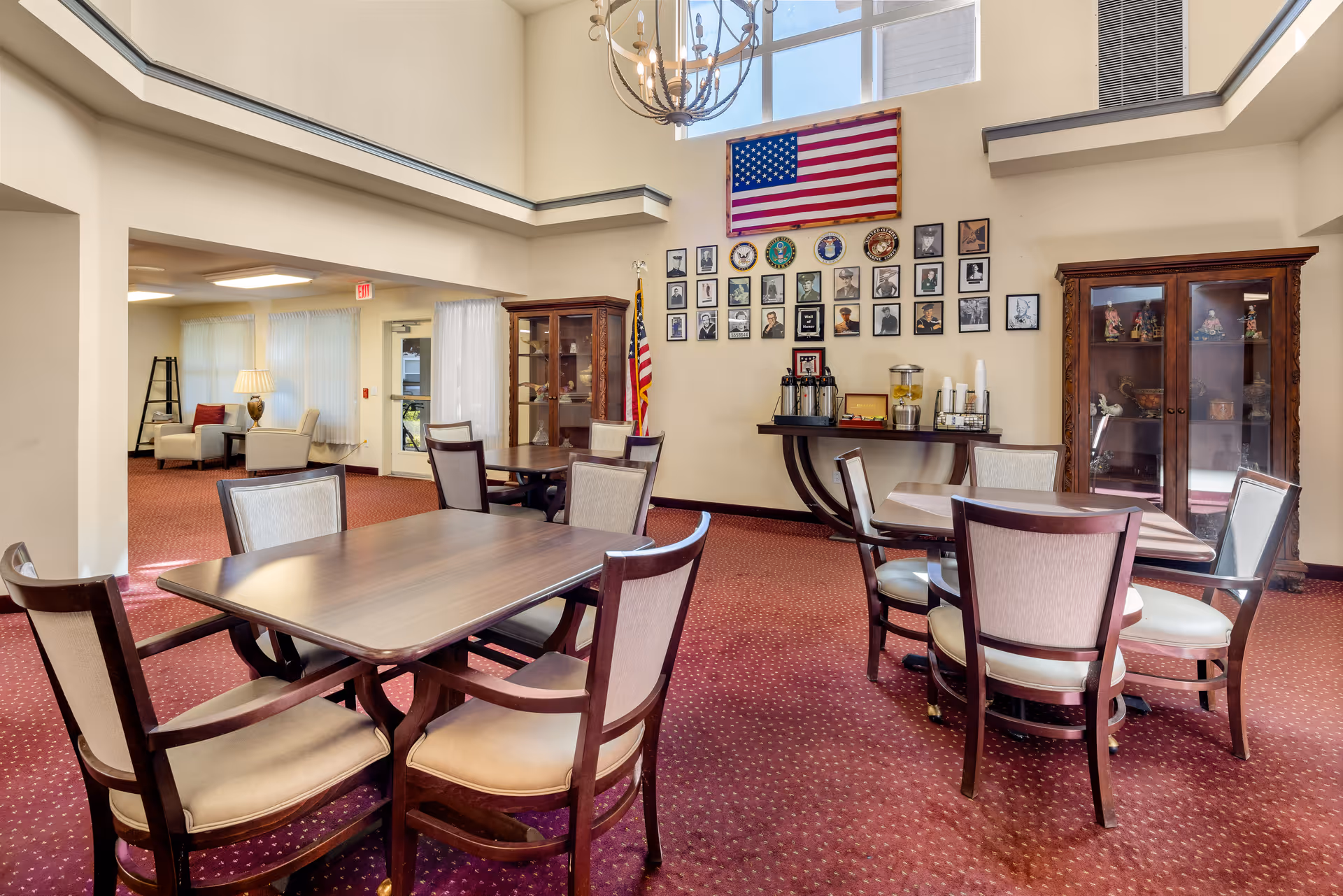 Interior of a senior living facility dining area with several wooden tables and cushioned chairs on a red carpet. The walls are decorated with an American flag, framed photographs, and military insignias. There are two wooden display cabinets and a refreshment table with coffee and water dispensers. Large windows allow natural light to fill the space.