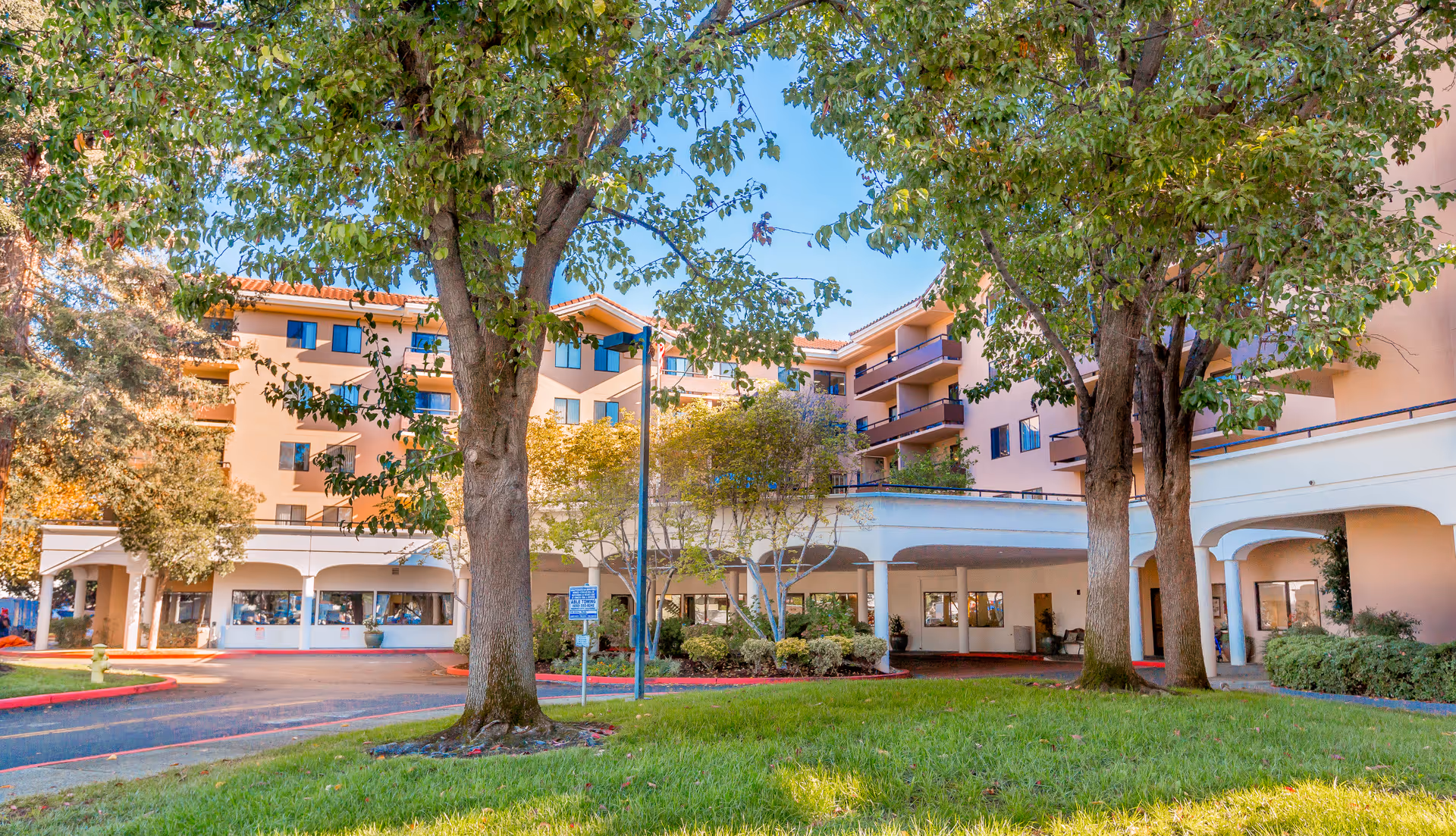 Exterior view of Brookdale Redwood City senior living facility showing a multi-story building with balconies, surrounded by trees and green lawn under a clear blue sky.