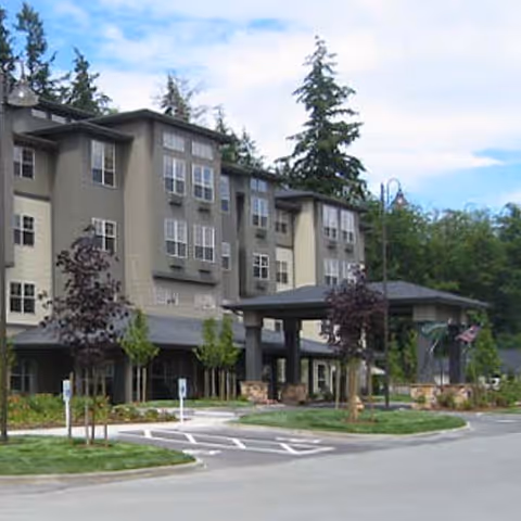 Exterior view of a multi-story senior living facility building with multiple windows, a covered entrance, landscaped greenery, and a driveway with parking spaces. Tall trees are visible in the background under a partly cloudy sky.
