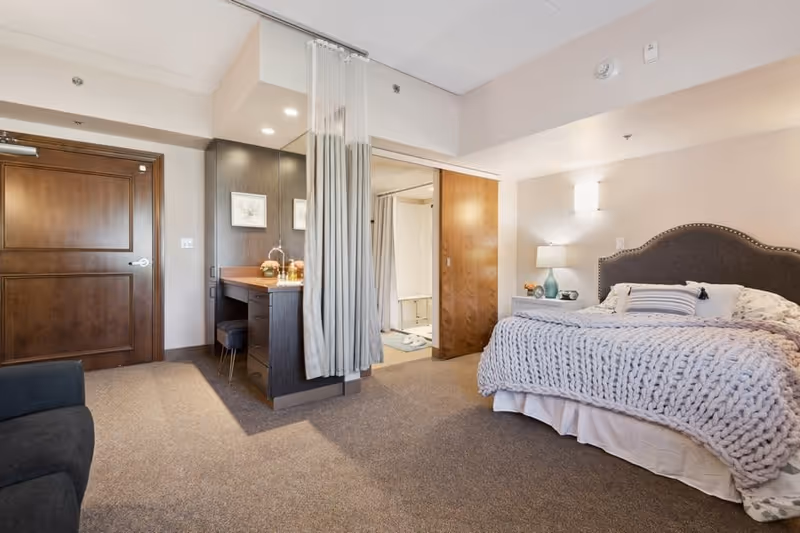 A cozy bedroom in a senior living facility featuring a large bed with a knitted blanket and pillows, a nightstand with a lamp and decorative items, a wooden door, a small vanity area with a stool and mirror, and a partially visible bathroom with a shower curtain.