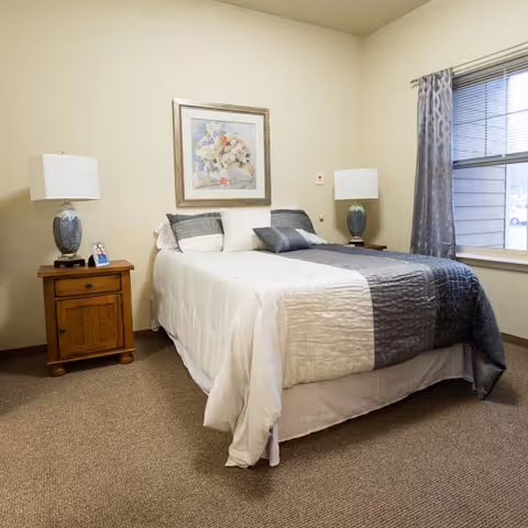 A neatly made bed with a white and gray comforter in a bedroom. There are two wooden nightstands on either side of the bed, each with a lamp. A framed floral painting hangs on the wall above the bed. A window with gray patterned curtains is on the right side of the room.