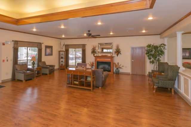A spacious living room area with wooden flooring, beige walls, and recessed ceiling lights. The room features several armchairs, a wooden coffee table with shelves, a fireplace with decorative plants and a mirror above it, windows with curtains, and a ceiling fan. There are also some framed pictures on the walls and a potted plant near a wooden door.
