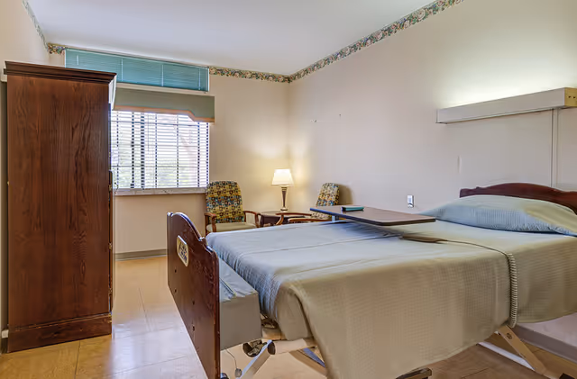 A simple hospital-style bedroom with a single bed covered in a beige blanket, a wooden wardrobe, two patterned chairs, a small table with a lamp, and a window with blinds and a green valance.