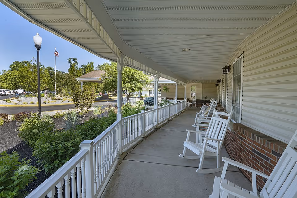 Covered porch area at Mattison Crossing with white rocking chairs lined up along a white railing. The porch overlooks a landscaped garden with bushes and trees, and a parking area with cars is visible in the background under a clear blue sky.