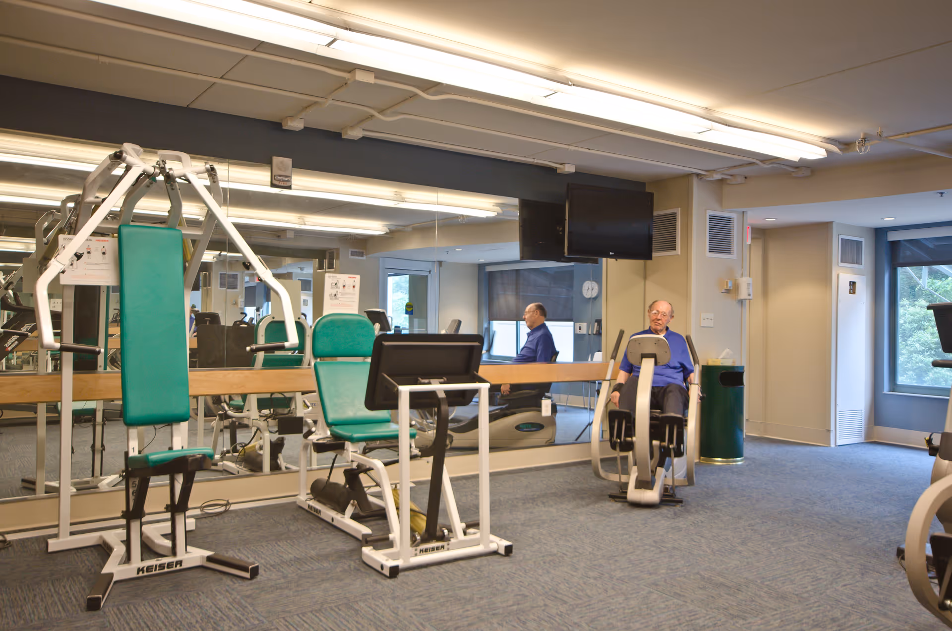 An elderly man exercising on a leg press machine in a fitness room with various exercise equipment, large mirrors on the wall, and a window showing greenery outside.