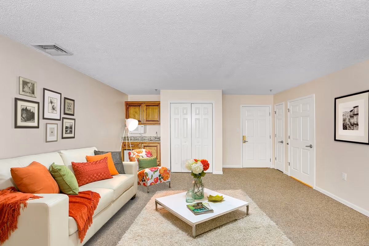 A cozy living room with a white sofa adorned with orange, red, and green pillows and a red throw blanket. There is a floral-patterned armchair next to a floor lamp. A white square coffee table with a vase of flowers, a small green dish, and magazines sits on a beige shag rug. The walls are decorated with framed black and white artwork. In the background, there is a small kitchenette with wooden cabinets and a granite countertop, and several white doors along the far wall.
