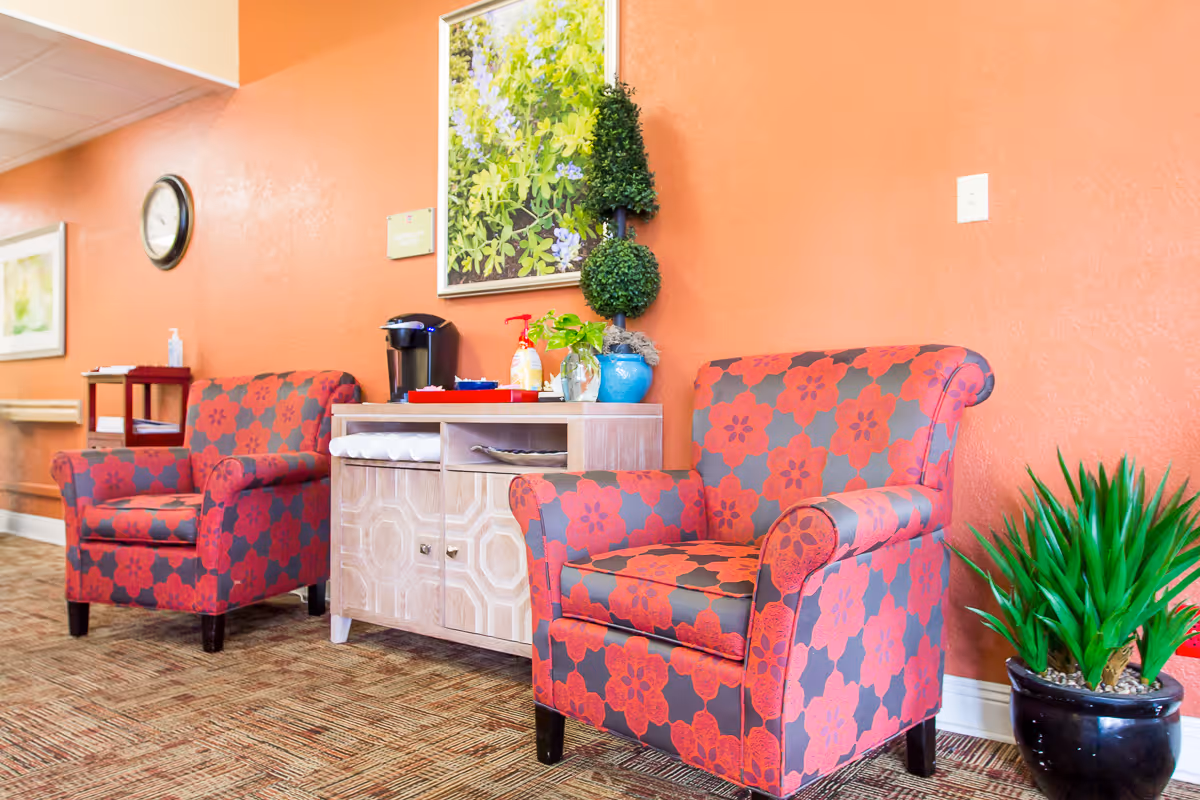 A cozy seating area with two red and gray floral patterned armchairs placed against an orange wall. Between the chairs is a light wooden cabinet with a coffee maker, hand sanitizer, and decorative plants on top. A framed picture of green foliage hangs on the wall above the cabinet. There is also a green potted plant on the floor to the right of the rightmost chair.
