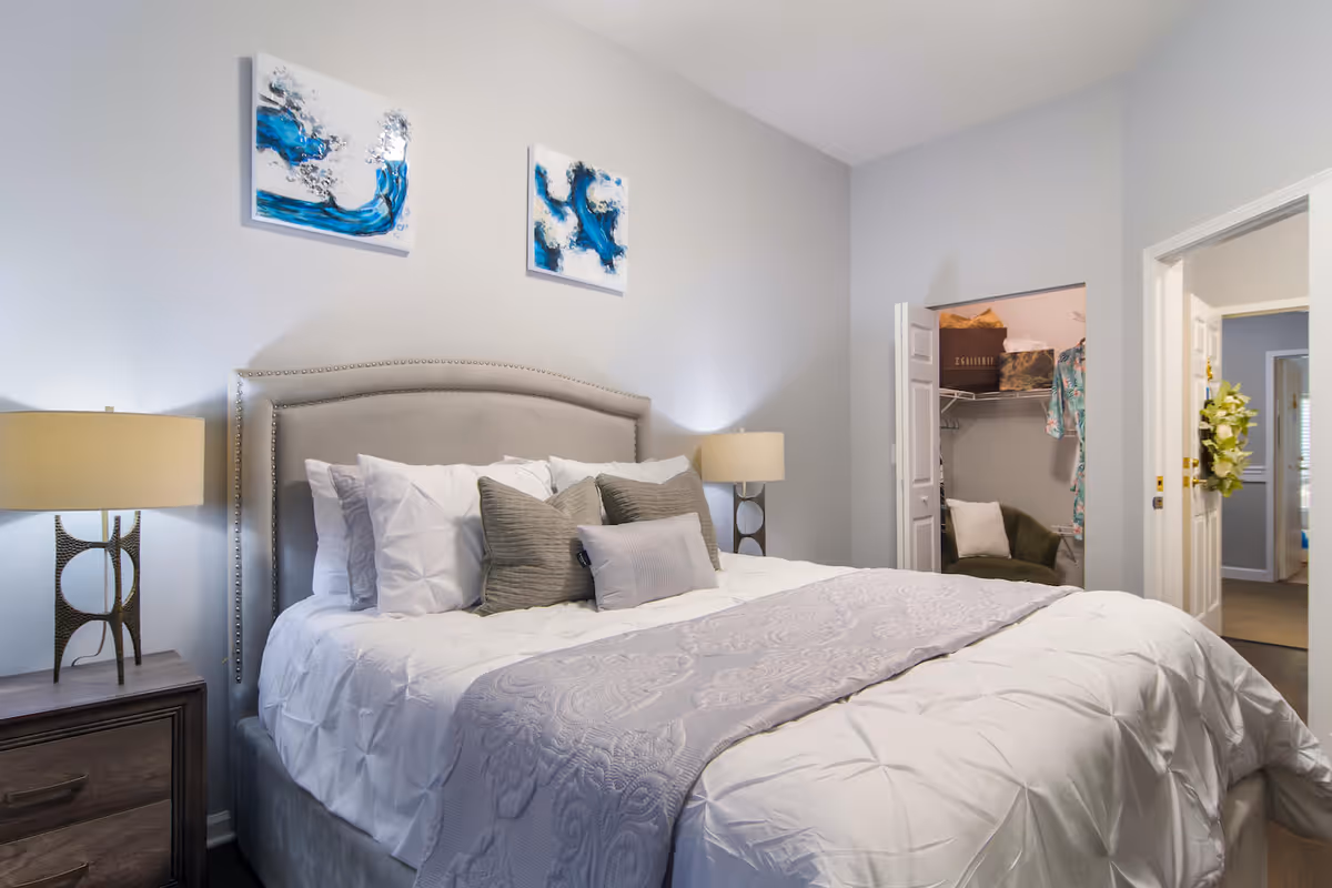 A neatly made bed with white and gray bedding in a bedroom. The bed has a padded headboard with nailhead trim. There are two matching bedside tables with modern lamps on each side. Above the bed, two abstract blue and white paintings hang on the wall. To the right, an open door reveals a walk-in closet with a chair and some hanging clothes. Another open door leads to an adjacent room.