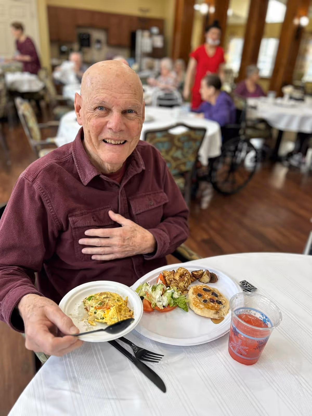 An older man smiles at the camera while sitting at a dining table with plates of breakfast and a drink, with other residents and tables in the background.