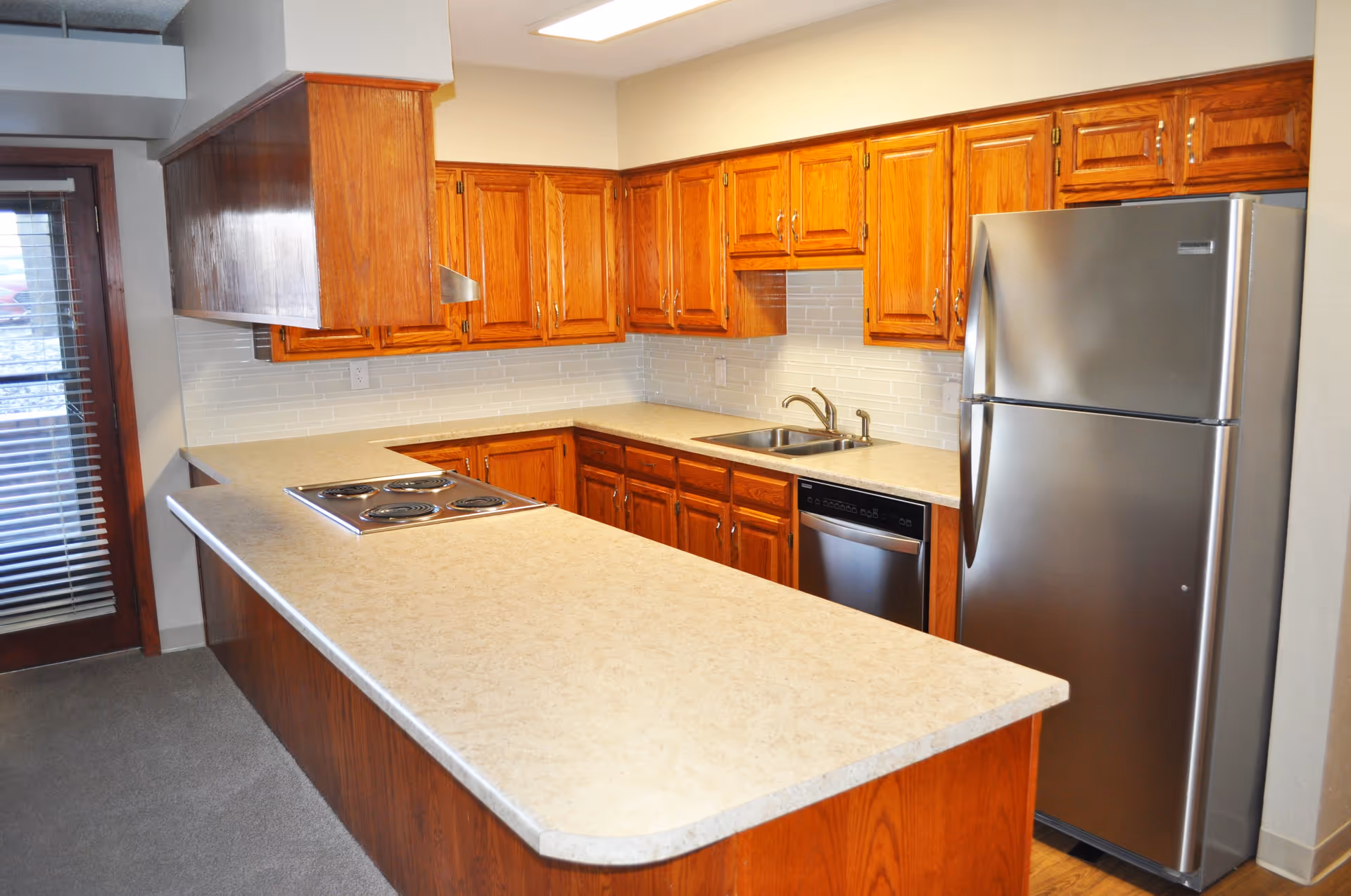Kitchen area with wooden cabinets, a beige countertop, a stainless steel refrigerator, a built-in electric stovetop, a dishwasher, and a sink. There is a door with blinds on the left side.