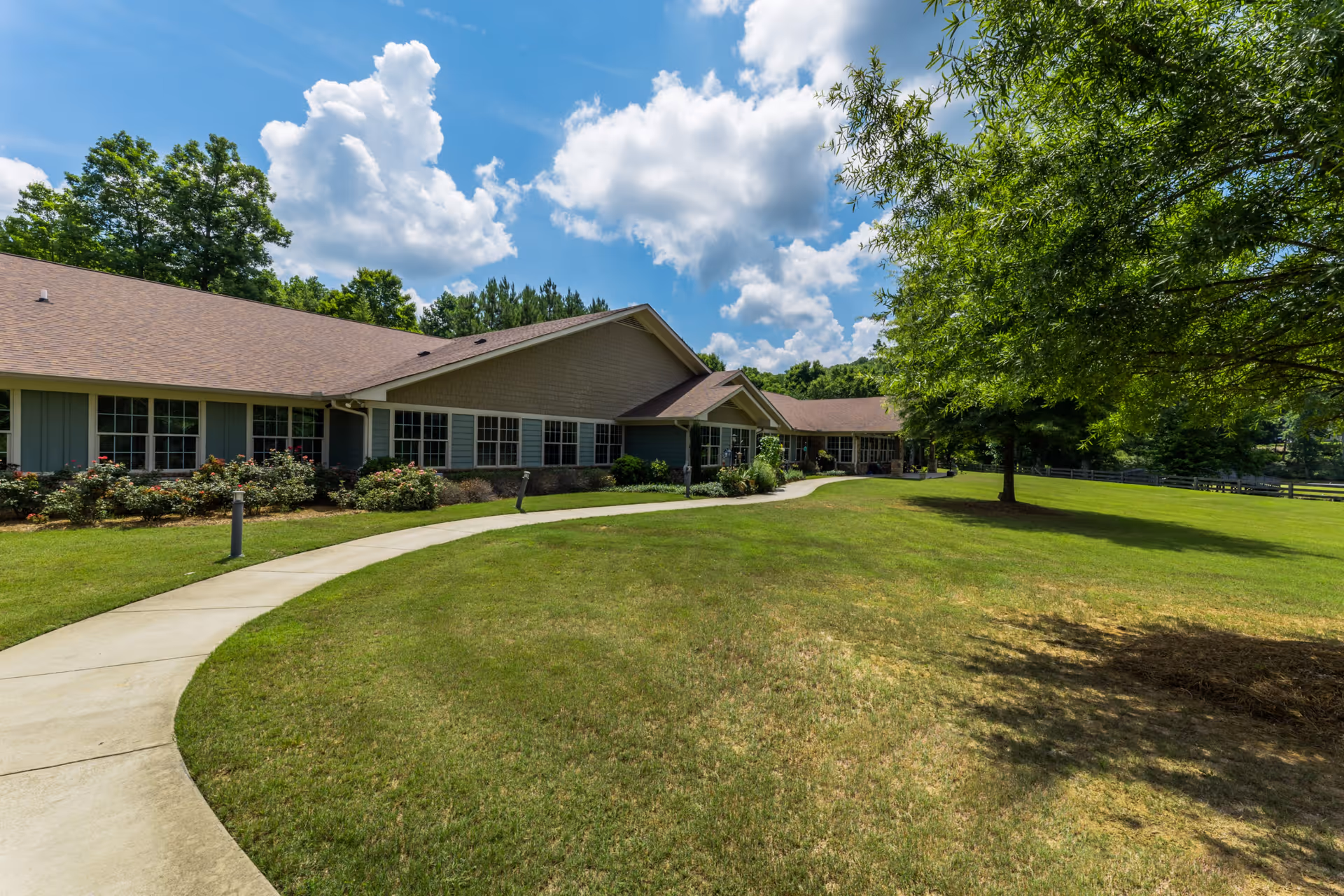Exterior view of Cedarhurst Senior Living of Canton showing a single-story building with multiple windows, surrounded by a well-maintained lawn, trees, and a curved concrete walkway under a partly cloudy blue sky.
