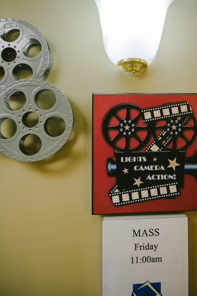 Wall decoration featuring two large silver film reels, a red sign with a black film camera and the words 'Lights Camera Action!', and a notice below announcing a Mass on Friday at 11:00 am.