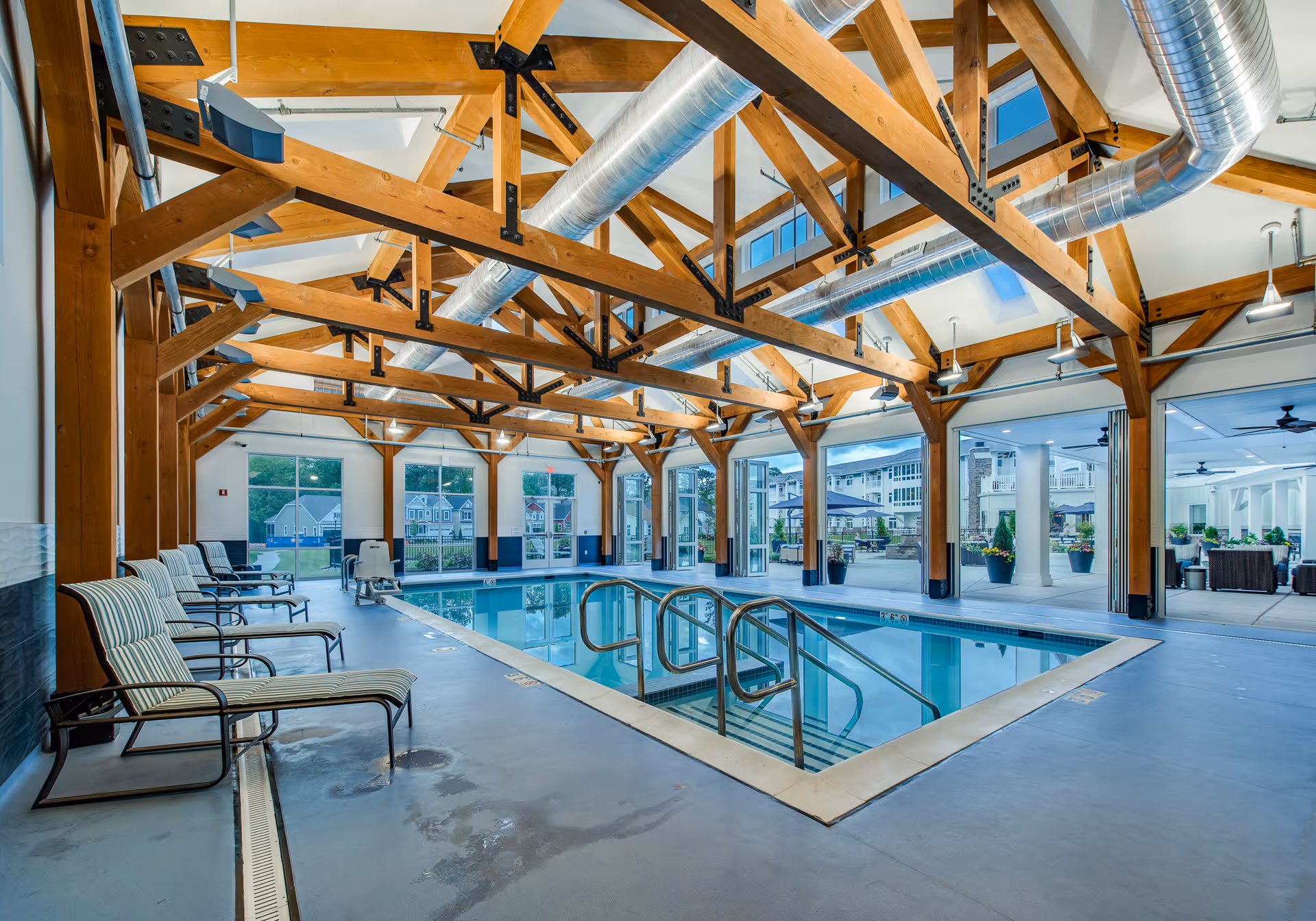 Indoor community swimming pool with striped lounge chairs, exposed wooden trusses, and large windows opening to an outdoor courtyard.