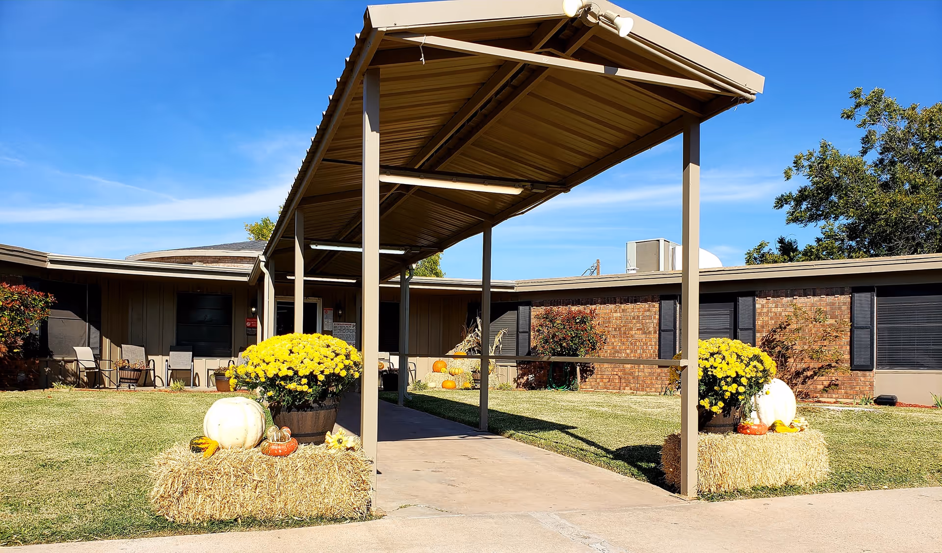 Outdoor view of a nursing and rehabilitation facility entrance with a covered walkway. The area is decorated with yellow flowers, pumpkins, and hay bales on a sunny day with a clear blue sky. The building has brick and beige siding with several windows and some outdoor chairs visible.