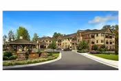 Exterior view of a large, multi-story senior living facility with a driveway leading to the entrance. The building is surrounded by landscaped greenery, trees, and a stone gazebo structure near the driveway. The sky is clear with a few clouds.