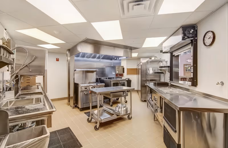 A clean, well-lit commercial kitchen with stainless steel appliances and countertops. The kitchen includes a large stove with an overhead vent, a metal prep table on wheels, multiple sinks, shelving units, and a clock on the wall. The floor is tiled and the ceiling has recessed lighting panels.