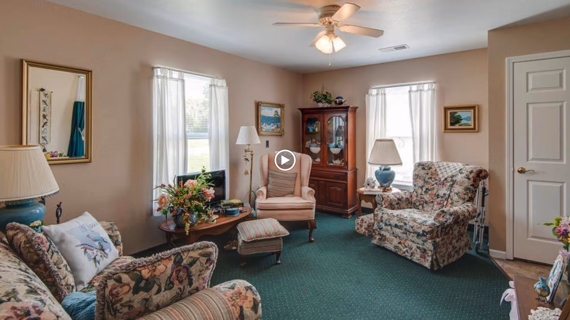 Cozy living room with floral-upholstered chairs and sofa, a coffee table with flowers, a china cabinet, and windows with sheer curtains.