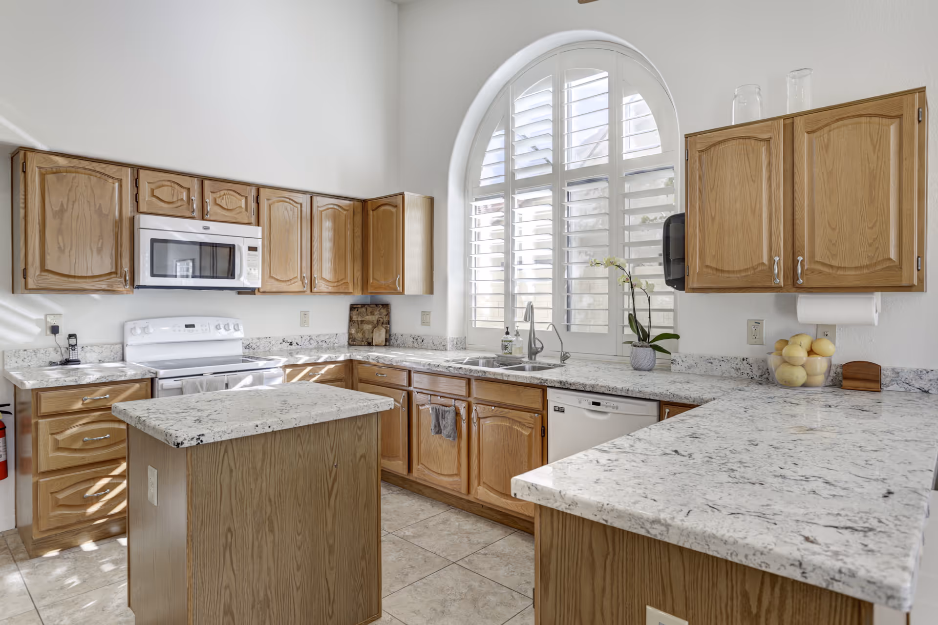 Bright kitchen with wooden cabinets, white granite countertops, a central island, a white stove with a microwave above it, a dishwasher, and a large arched window with white shutters letting in natural light.