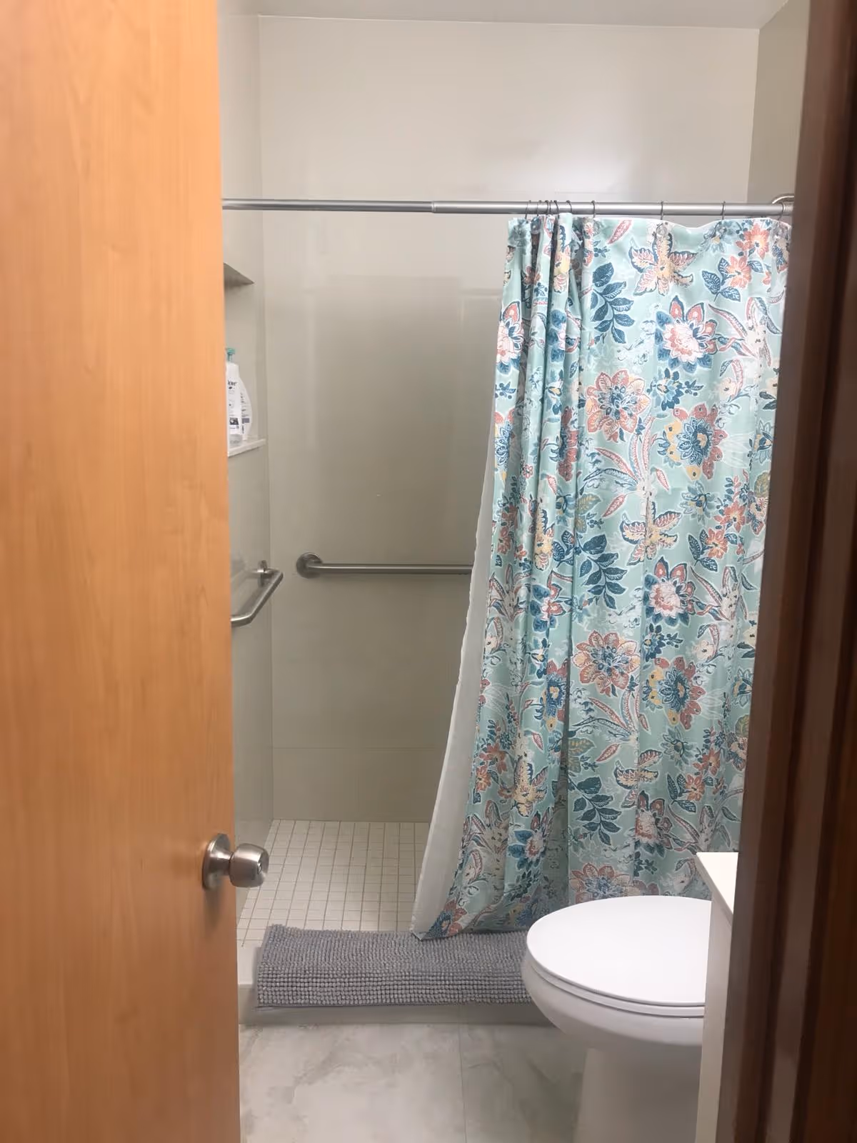 View into a bathroom with a partially open wooden door. Inside, there is a toilet on the right and a shower area with a floral patterned shower curtain. The shower has a grab bar on the wall and a gray bath mat on the floor outside the shower.