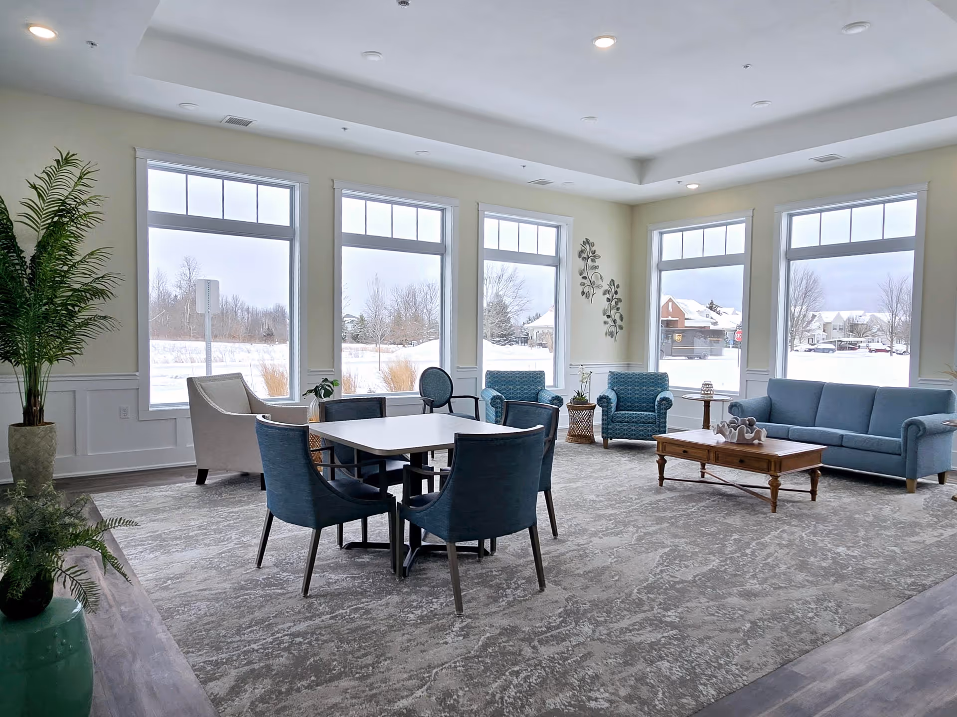 A bright and spacious living room area with large windows showing a snowy outdoor scene. The room features a blue sofa, two blue patterned armchairs, a wooden coffee table with decorative items, a small round side table, a square table with four blue chairs, and a beige armchair. There are plants and wall decorations adding to the cozy atmosphere.
