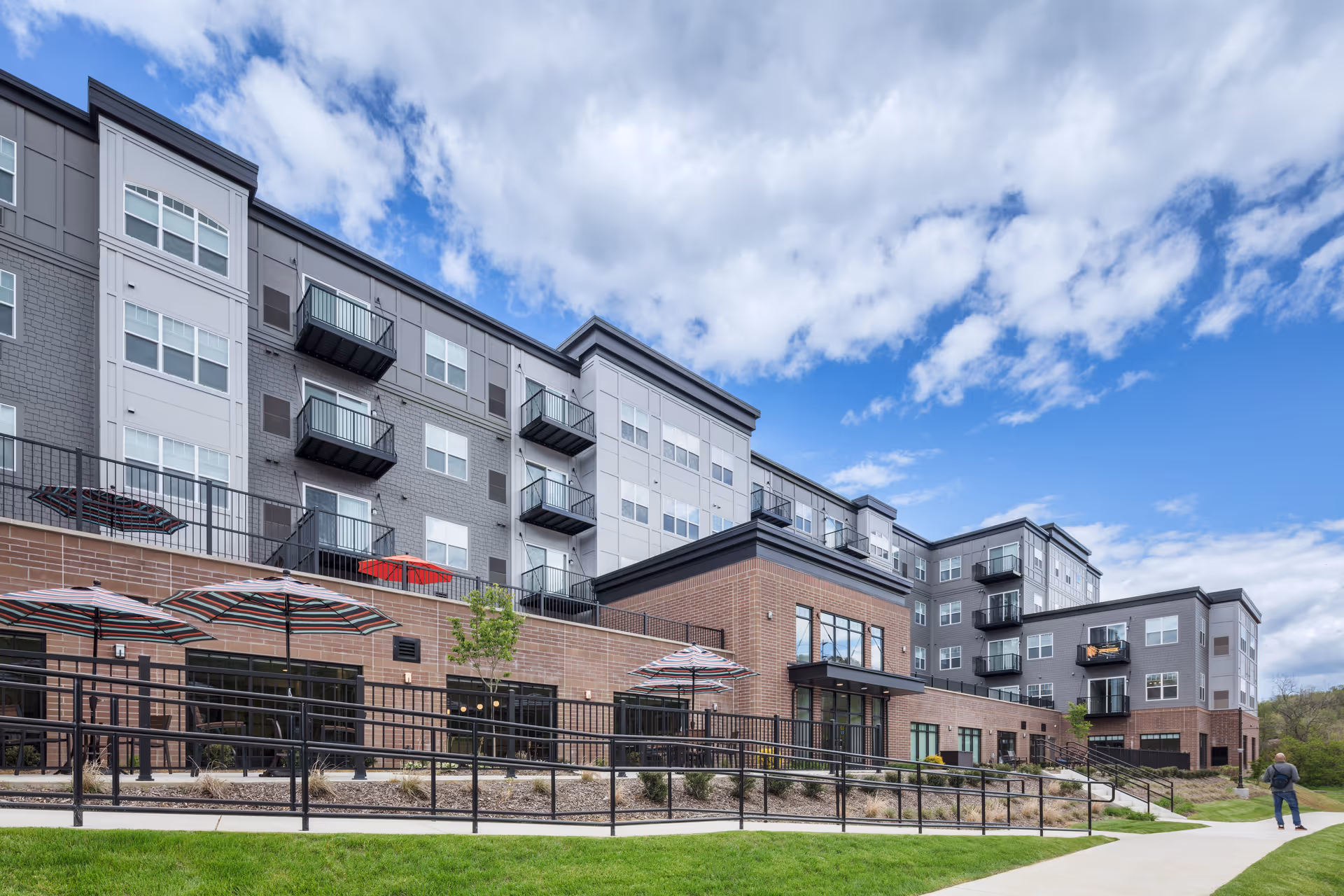Exterior view of a modern multi-story senior living facility with balconies, large windows, and outdoor patio umbrellas. The building has a combination of brick and gray siding under a partly cloudy blue sky. A person is walking on the sidewalk near the building.