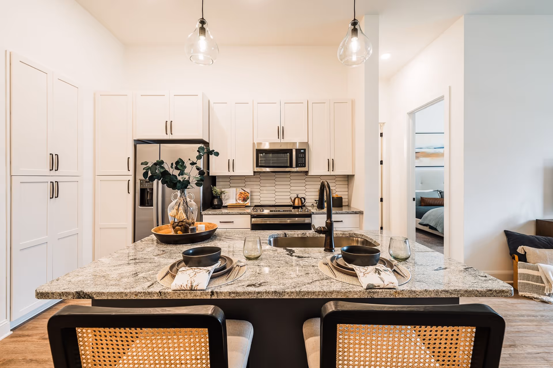 Modern kitchen with a large granite island countertop set for two with black bowls, plates, and glasses. The kitchen features white cabinets, stainless steel refrigerator, oven, and microwave. Two pendant lights hang above the island. A doorway leads to a bedroom visible in the background.
