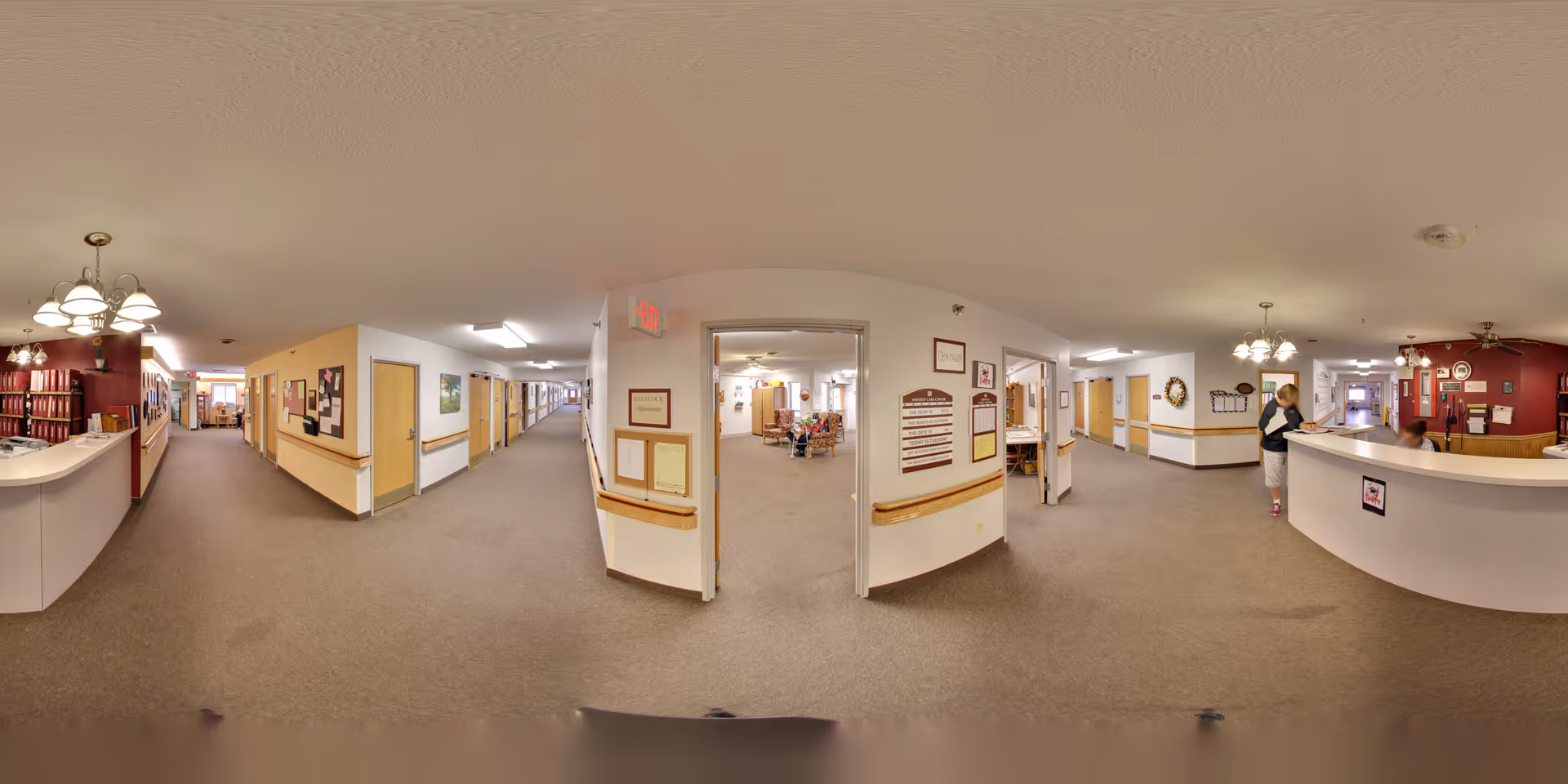 Panoramic interior of a senior living facility hallway showing reception desks on both sides, a central doorway to a common area, and long corridors.