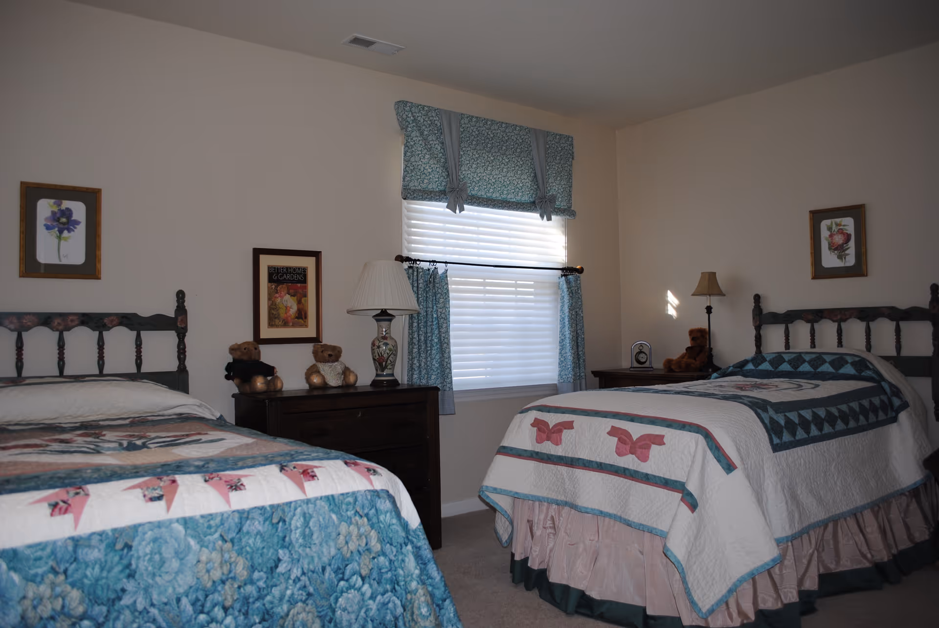 A bedroom with two twin beds, each with floral and butterfly patterned quilts. Between the beds is a window with blue floral curtains and a valance. There are two dark wooden nightstands, each with a lamp and teddy bears. The walls are decorated with framed floral artwork.